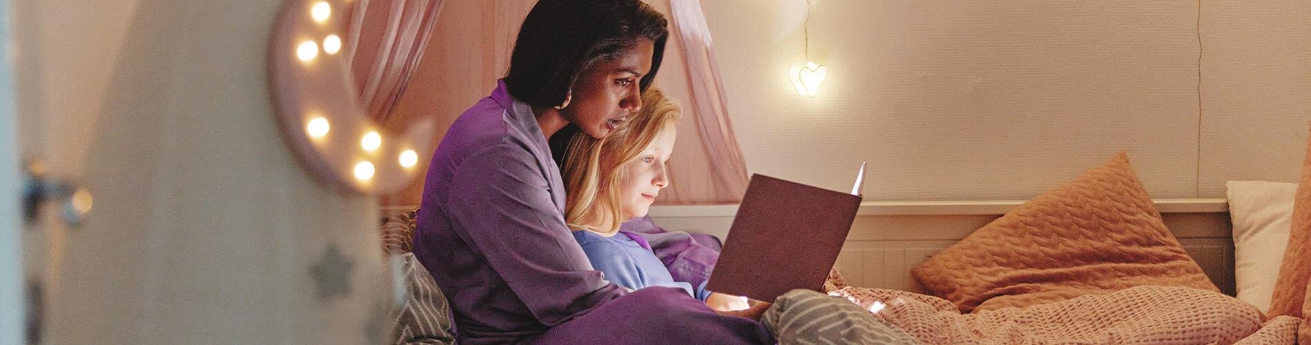 A woman reading a book with a child while sitting on a bed. 